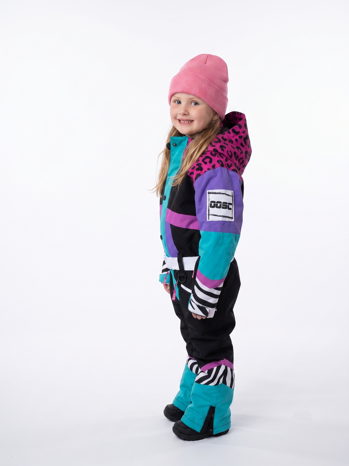 A young girl modeling a colorful winter ski suit with a pink beanie, standing against a plain white studio background. The ski suit features a color-blocked design of black, purple, teal, and pink with leopard and zebra print details.
